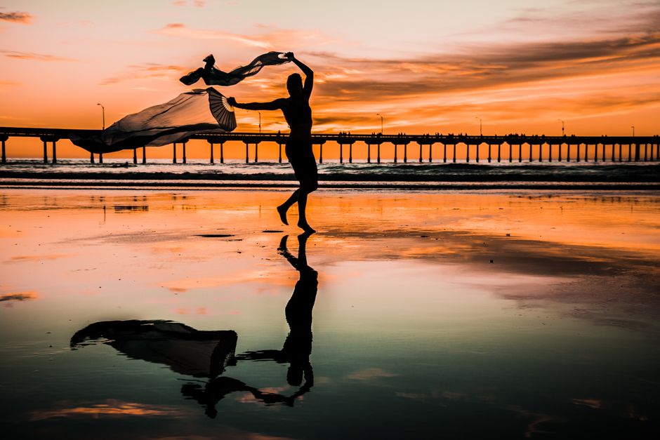 A graceful figure dances with a scarf during sunset at the beach, creating a stunning silhouette reflection.